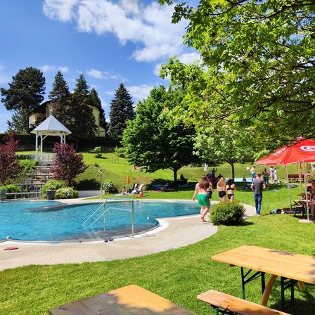Ein sonniger Tag an einem Außenpool mit Sonnenbadenden und Spaziergängern, umgeben von Bäumen, einem Pavillon und Picknicktischen.