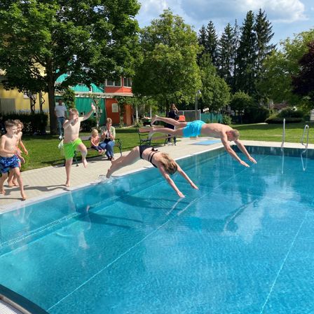Mehrere Kinder springen an einem sonnigen Tag in ein Schwimmbecken, während Zuschauer von der Seite zusehen. Einige Erwachsene sind ebenfalls anwesend und sitzen auf einer Bank. Das Becken ist von einer Rasenfläche und Bäumen umgeben.