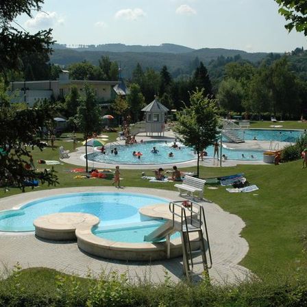 Freibad mit mehreren Schwimmenden, einer Wasserrutsche für Erwachsene und einer kleinen Wasserrutsche. Menschen entspannen sich auf der Wiese und einige sitzen auf Bänken.