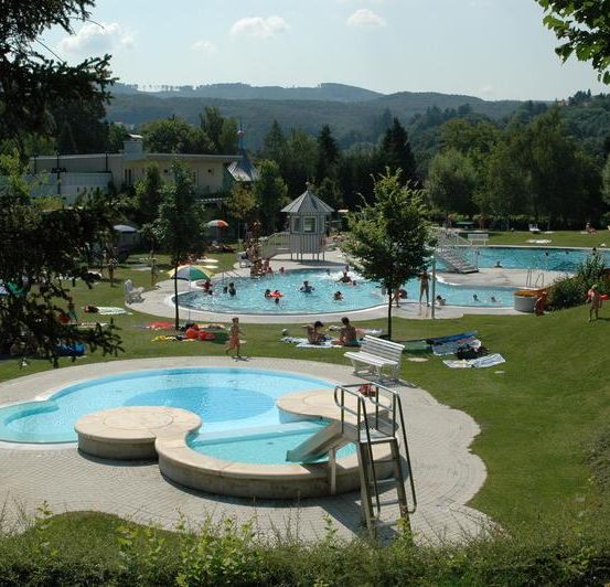 Freibad mit mehreren Schwimmenden, einer Wasserrutsche für Erwachsene und einer kleinen Wasserrutsche. Menschen entspannen sich auf der Wiese und einige sitzen auf Bänken.
