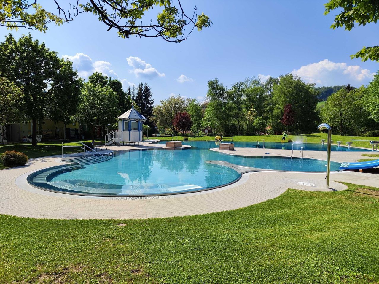 Ein Freibad mit einem weißen und blauen Pavillon. Bäume und Pflanzen umgeben den Pool. Eine Person steht an der Seite des Pools.