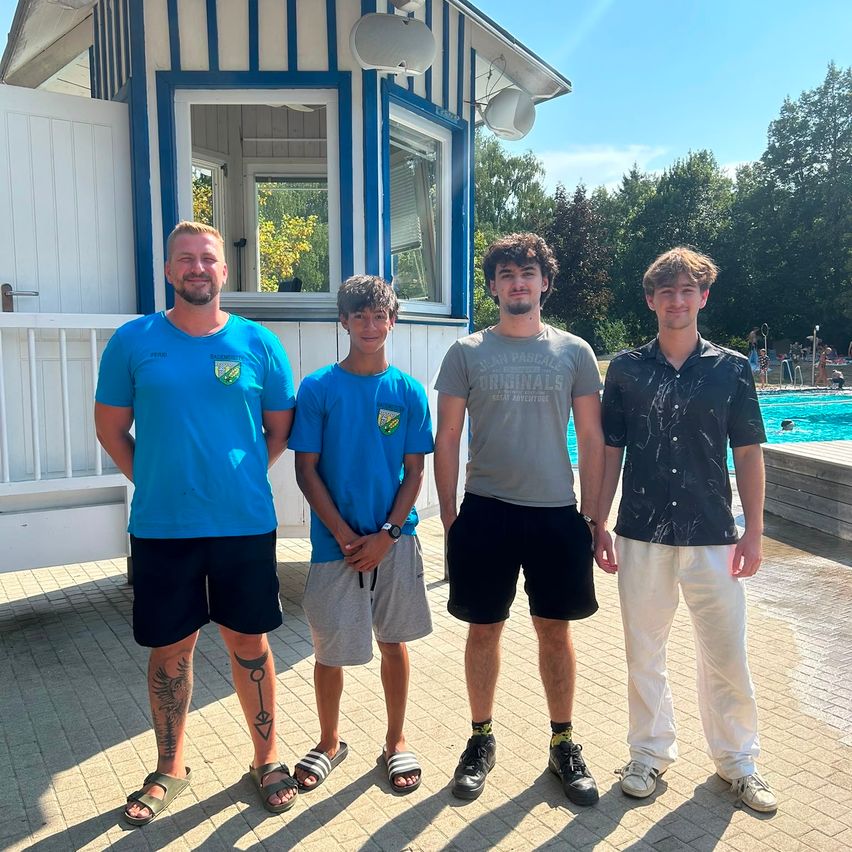 Vier Männer stehen vor einem Gebäude mit einem Schwimmbad im Hintergrund. Sie tragen alle Freizeitkleidung.