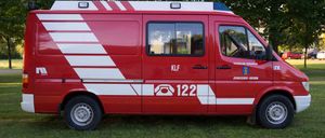 Ein roter Van mit weißen Streifen steht auf einem Rasenfeld. Er hat den Text 'KLF' und '122' an der Seite und ein Logo an der Tür.