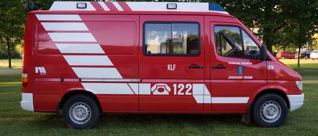 Ein roter Van mit weißen Streifen steht auf einem Rasenfeld. Er hat den Text 'KLF' und '122' an der Seite und ein Logo an der Tür.