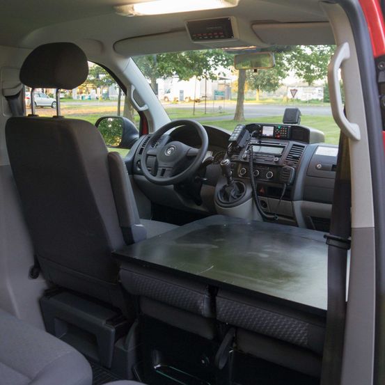 Innenraum eines Fahrzeugs mit Fahrersitz, Lenkrad, Armaturenbrett und Rückspiegel. Draußen vor dem Fenster ein Rasen mit Bäumen und einer Straße.