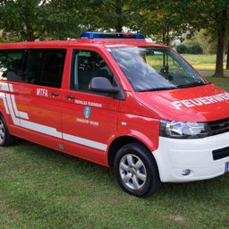 Ein roter Feuerwehrwagen ist auf grünem Gras mit Bäumen im Hintergrund geparkt. Es steht 'FEUERWEHR' an der Seite.