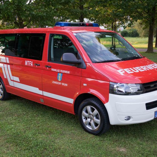 Ein roter Feuerwehrwagen ist auf grünem Gras mit Bäumen im Hintergrund geparkt. Es steht 'FEUERWEHR' an der Seite.