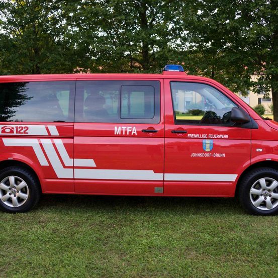 Ein roter Feuerwehrwagen mit MTFA und Freiwillige Feuerwehr Johnsdorf-Brunn Aufschriften, geparkt auf einem Rasenfeld mit Bäumen im Hintergrund.