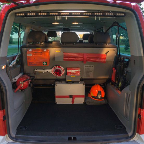 Innenraum eines roten Vans mit Notfallausrüstung, einschließlich Feuerlöscher, Erste-Hilfe-Kasten und Helm. Die Rücksitze sind umgeklappt und im Laderaum befinden sich ein Werkzeugkasten, eine Aufbewahrungsbox und weitere Werkzeuge.