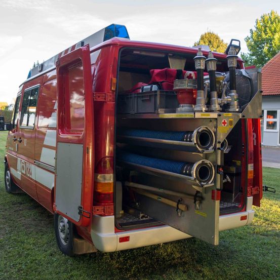 Ein roter Feuerwehrwagen mit offenen Hecktüren steht auf einem Rasenfeld. Im Inneren befinden sich Lagerboxen, Feuerlöscher und Schläuche. Im Hintergrund ist ein Haus mit einem roten Dach zu sehen.