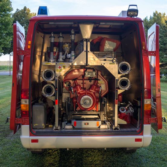 Die Rückseite eines roten Feuerwehrwagens mit offenen Hecktüren, die eine Feuerpumpe, Düsen und Ausrüstung auf dem Rasen zeigt. Bäume und ein Gebäude sind im Hintergrund.