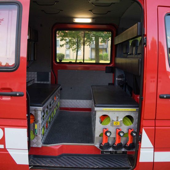 Die Rückseite eines roten Notfallfahrzeugs, offen. Im Inneren befinden sich zwei Abteile mit verschiedenen Werkzeugen und Ausrüstungsgegenständen. Ein Fenster zeigt ein Gebäude und Bäume draußen.