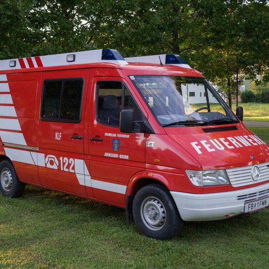 Ein roter Feuerwehrwagen mit weißen Streifen ist auf einem Rasen geparkt. Der Wagen hat das Wort 'FEUERWEHR' an der Seite.