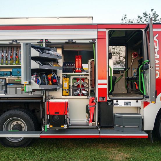 Ein rot-weißer Feuerwehrwagen mit offenen Türen, der einen organisierten Innenraum mit verschiedenen Werkzeugen und Ausrüstung zeigt.