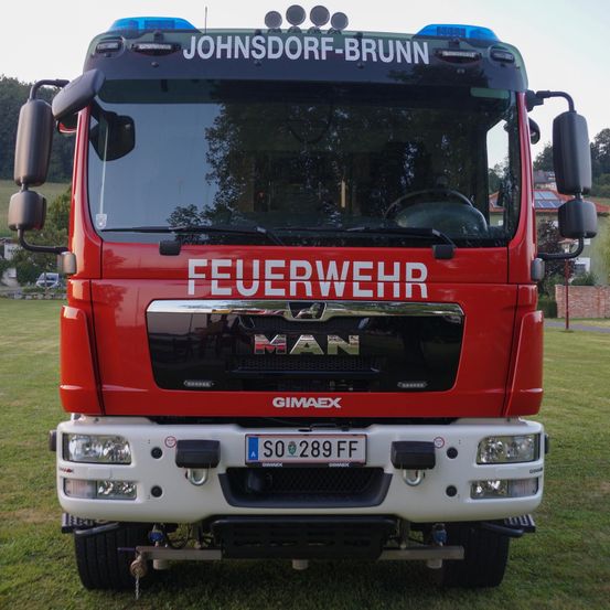 Ein roter Feuerwehrwagen mit 'Johnsdorf-Brunn' oben und 'Feuerwehr' vorne ist auf einem grünen Feld geparkt. Das Kennzeichen lautet 'SO 289 FF'.