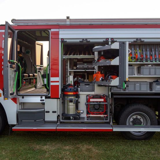 Die Rückseite eines roten und weißen Feuerwehrwagens mit offenen Türen. Im Inneren sind Werkzeuge und Ausrüstung auf Regalen und in Behältern angeordnet.