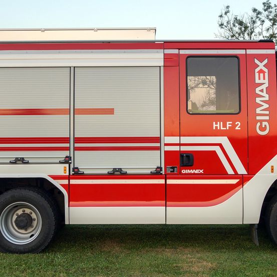 Ein rot-weißes Notfallfahrzeug, gekennzeichnet mit 'HLF 2' und 'GIMAEX', auf grünem Gras geparkt.