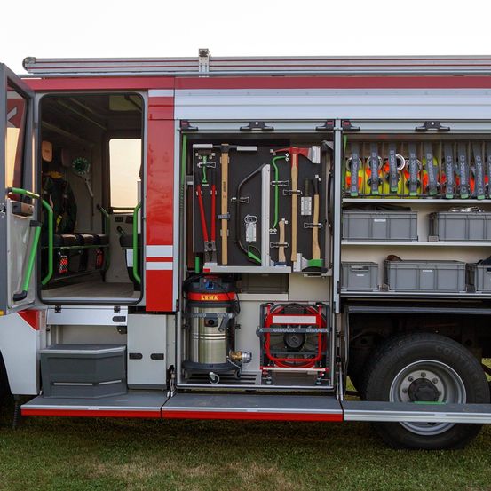 Die Rückseite eines Feuerwehrautos mit offenen Türen, die verschiedene Werkzeuge und Ausrüstung im Inneren zeigen. Sichtbare Aufbewahrungsfächer und -boxen, in der Mitte ein silberner Staubsauger.