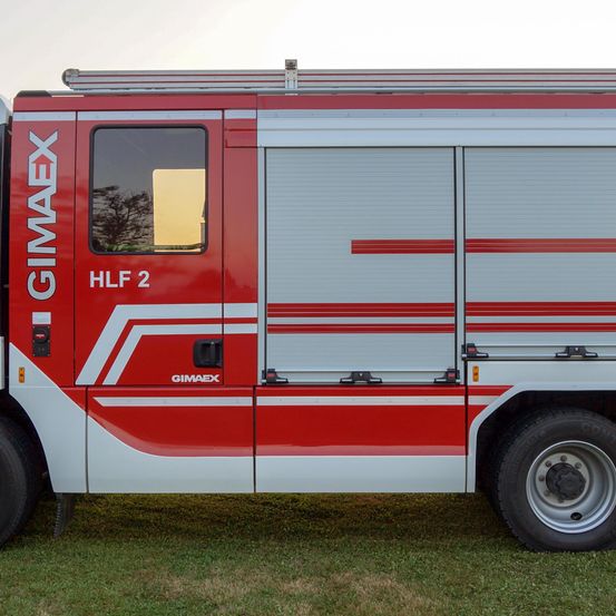 Ein rot-weißer Feuerwehrwagen mit 'GIMAEX' und 'HLF 2' steht auf grünem Gras.