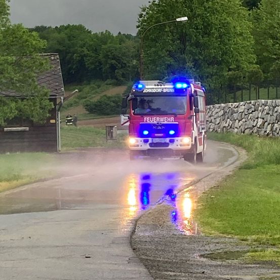 Ein roter Feuerwehrwagen mit blauen Lichtern fährt auf einer nassen Straße, mit einem Haus und Bäumen im Hintergrund.