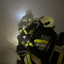 Drei Feuerwehrleute in voller Ausrüstung, mit Helmen, befinden sich in einem rauchigen Bereich. Sie sind mit Taschenlampen und Gurten ausgestattet. Der in der Mitte hält eine Maske.