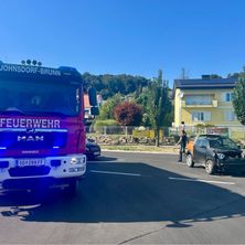 Ein Feuerwehrwagen aus Johnsdorf-Brunn mit blinkenden Lichtern ist auf einer Straße geparkt. Eine Person steht neben einem demolierten Auto. Im Hintergrund ist ein gelbes Haus mit Solarmodulen zu sehen.