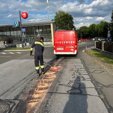 Ein Feuerwehrmann reinigt die Straße vor einem Bahnhof. Ein roter Van mit dem Wort 'Feuerwehr' ist in der Nähe geparkt. Bäume und ein Gebäude im Hintergrund.