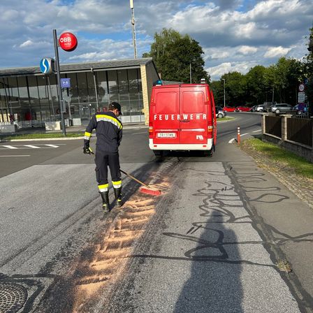 Ein Feuerwehrmann reinigt die Straße vor einem Bahnhof. Ein roter Van mit dem Wort 'Feuerwehr' ist in der Nähe geparkt. Bäume und ein Gebäude im Hintergrund.