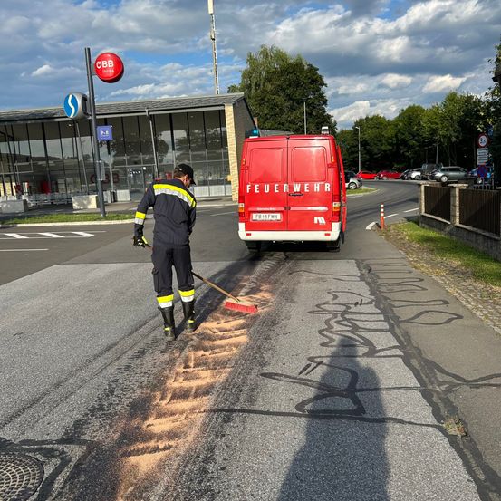 Ein Feuerwehrmann reinigt die Straße vor einem Bahnhof. Ein roter Van mit dem Wort 'Feuerwehr' ist in der Nähe geparkt. Bäume und ein Gebäude im Hintergrund.