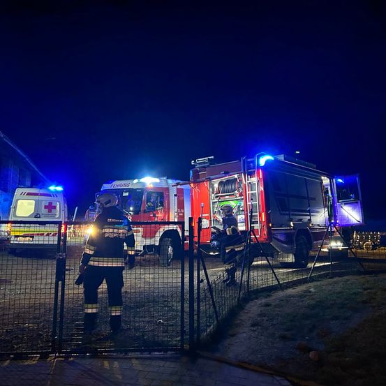 Zwei Feuerwehrleute stehen neben einem Feuerwehrauto mit blinkenden Lichtern. Ein Krankenwagen mit roten Lichtern ist in der Nähe geparkt. Es ist Nacht.