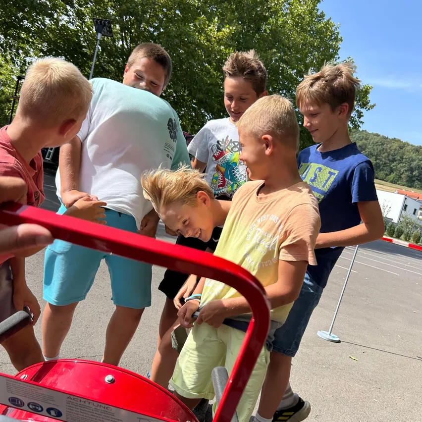 Mehrere Jungen sind um ein rotes Auto in einem Parkplatz versammelt, möglicherweise reparieren oder inspizieren sie es. Bäume und ein Gebäude sind im Hintergrund sichtbar.