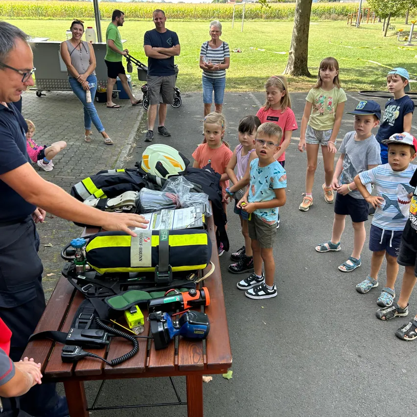 Eine Gruppe von Kindern und Erwachsenen versammelt sich um einen Tisch mit Sicherheitsausrüstung im Freien. Die Geräte umfassen Helme und Taschen. Erwachsene sind anwesend, einer schiebt einen Kinderwagen.