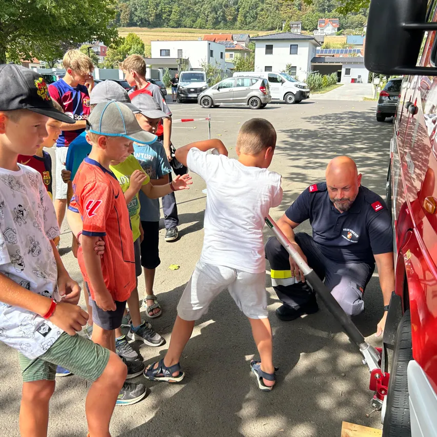 Ein Feuerwehrmann zeigt einer Gruppe von Kindern, wie man einen Feuerwehrwagenheber bedient. Sie stehen im Kreis um ihn herum und beobachten aufmerksam.