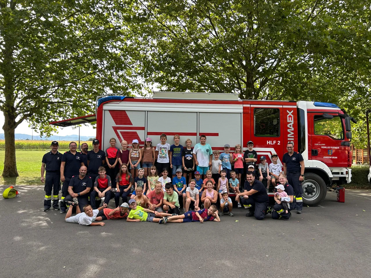 Eine Gruppe von Kindern und Erwachsenen posiert für ein Foto vor einem roten Feuerwehrauto. Einige sitzen auf dem Wagen, während andere stehen. Bäume sind im Hintergrund.