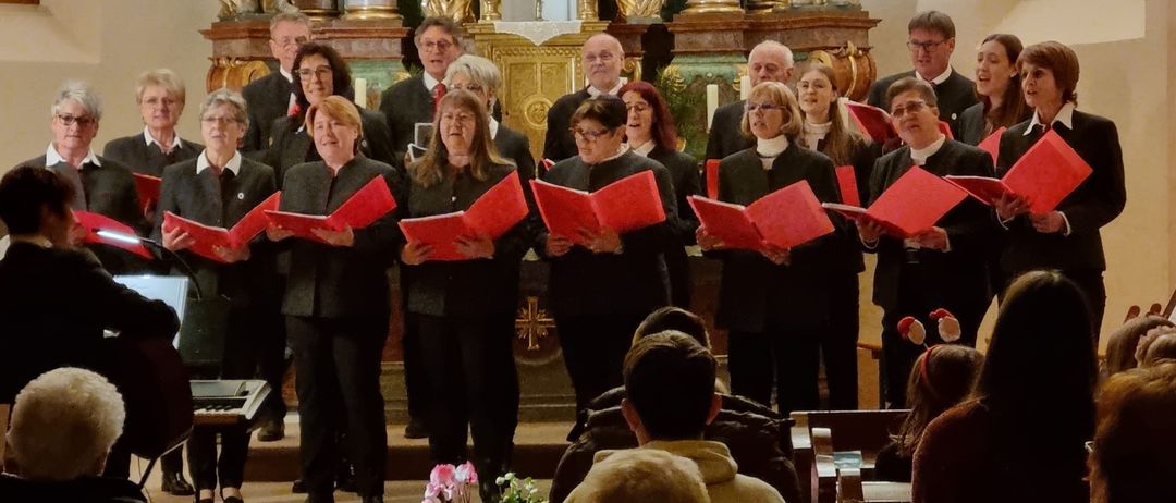 Ein Chor von Personen in schwarzer Kleidung steht vor einem Altar und hält rote Liederbücher in den Händen. Sie befinden sich in einer Kirche mit goldenen Statuen im Hintergrund. Das Publikum sitzt vorne und schaut auf den Chor.