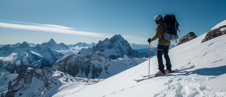 Ein Skifahrer steht auf einem verschneiten Berghang, ausgestattet mit Skiern, Stöcken und einem Helm. Der Skifahrer trägt eine grüne Jacke und einen Rucksack. Hinter ihm ist eine verschneite Bergkette.