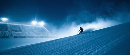 Ein Skifahrer fährt bei Nacht einen Hang hinab, beleuchtet von Flutlichtern. Der Skifahrer ist von Nebel umgeben, und das Stadion scheint leer zu sein.