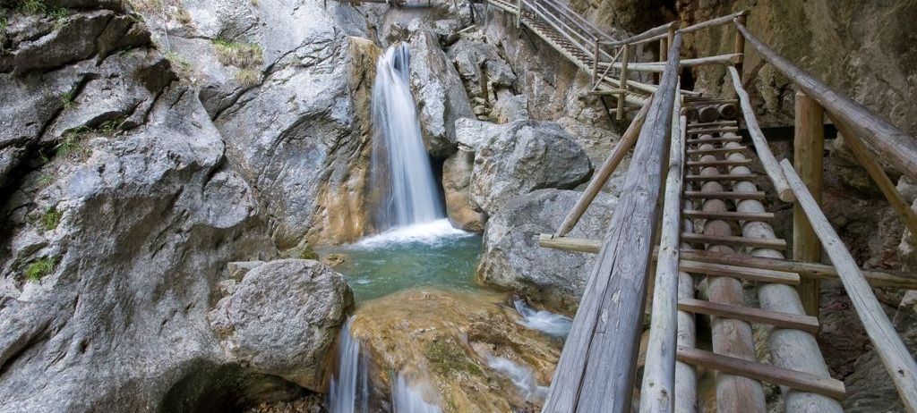Ein Wasserfall stürzt an einem felsigen Abhang herab, mit hölzernen Stufen und einem Handlauf, der zur Spitze führt.