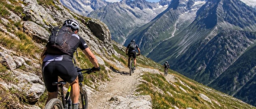 Drei Mountainbiker fahren auf einem rauen Pfad mit schneebedeckten Bergen im Hintergrund unter einem klaren blauen Himmel.