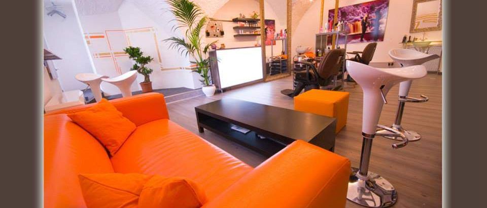 Ein Innenraum mit einer orangefarbenen Couch, einem Couchtisch, Hockern und Pflanzen. Im Hintergrund befinden sich ein Spiegel, Regale und ein Friseursalon. Das Bild hat einen Wasserzeichen mit einer Website-URL.