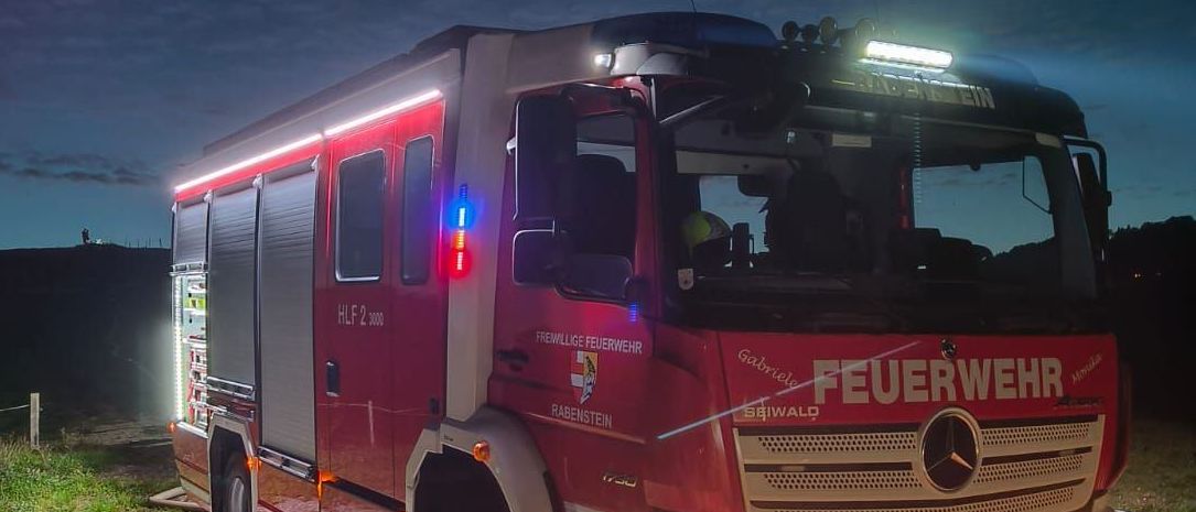 Ein roter Feuerwehrwagen mit eingeschalteten Scheinwerfern und Blaulicht steht nachts auf einer Straße geparkt.