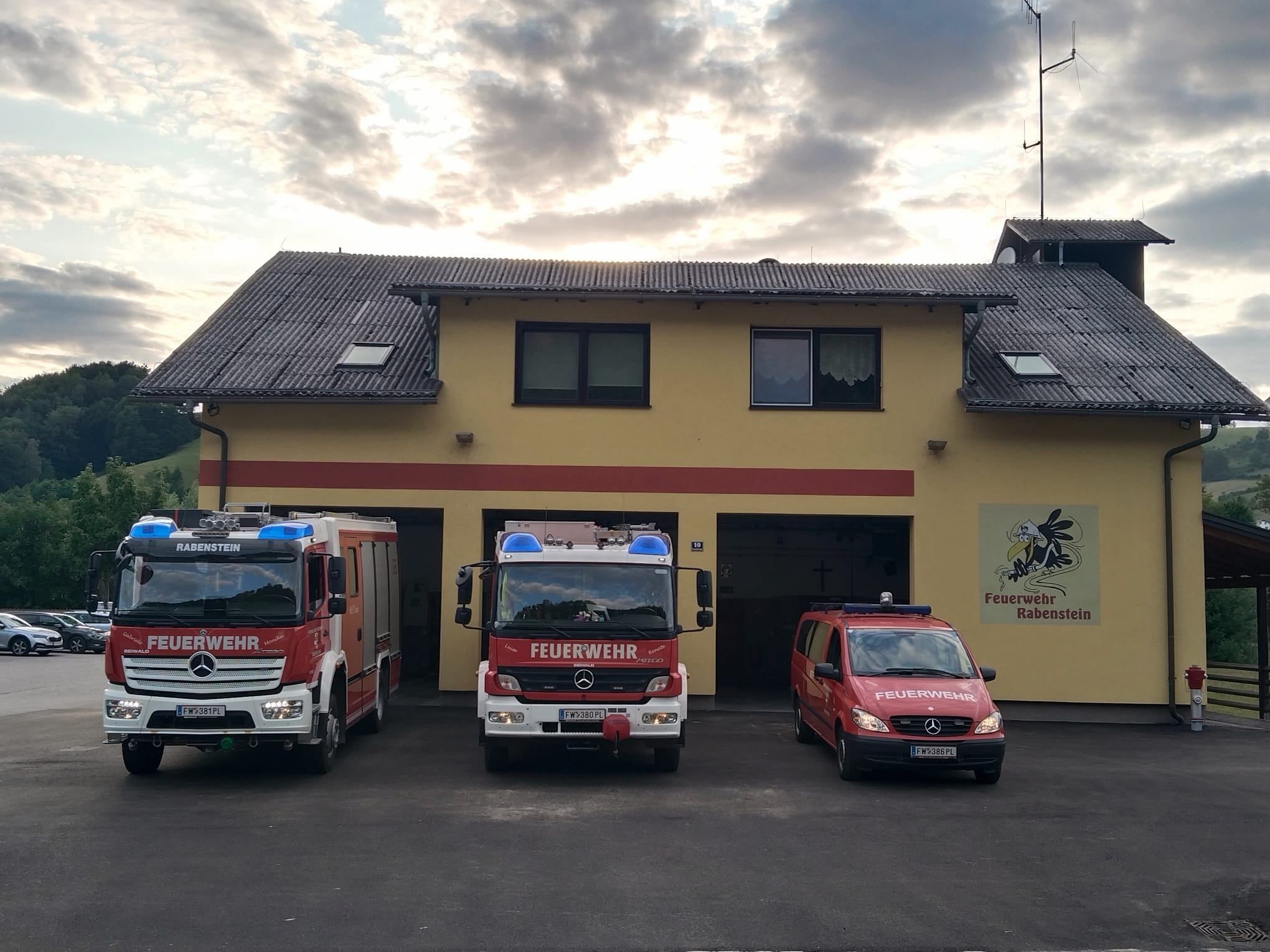 Drei Feuerwehrfahrzeuge sind vor einem Feuerwehrhaus mit einem Schild, das 'Feuerwehr Rabenstein' besagt, geparkt. Die Fahrzeuge sind rot mit weißen und blauen Lichtern auf der Oberseite. Der Himmel ist bewölkt.