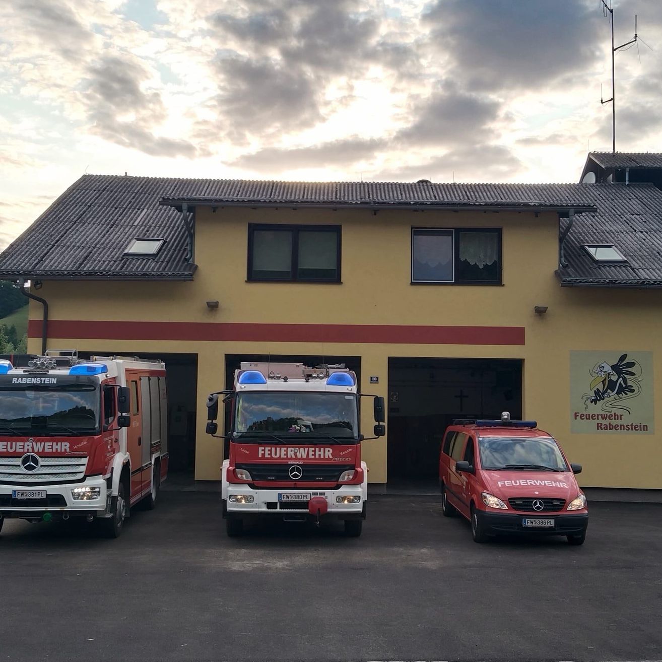 Drei Feuerwehrfahrzeuge stehen vor einem gelben Gebäude mit einem Schild, auf dem 'Feuerwehr Rabenstein' steht. Der Himmel ist bewölkt.