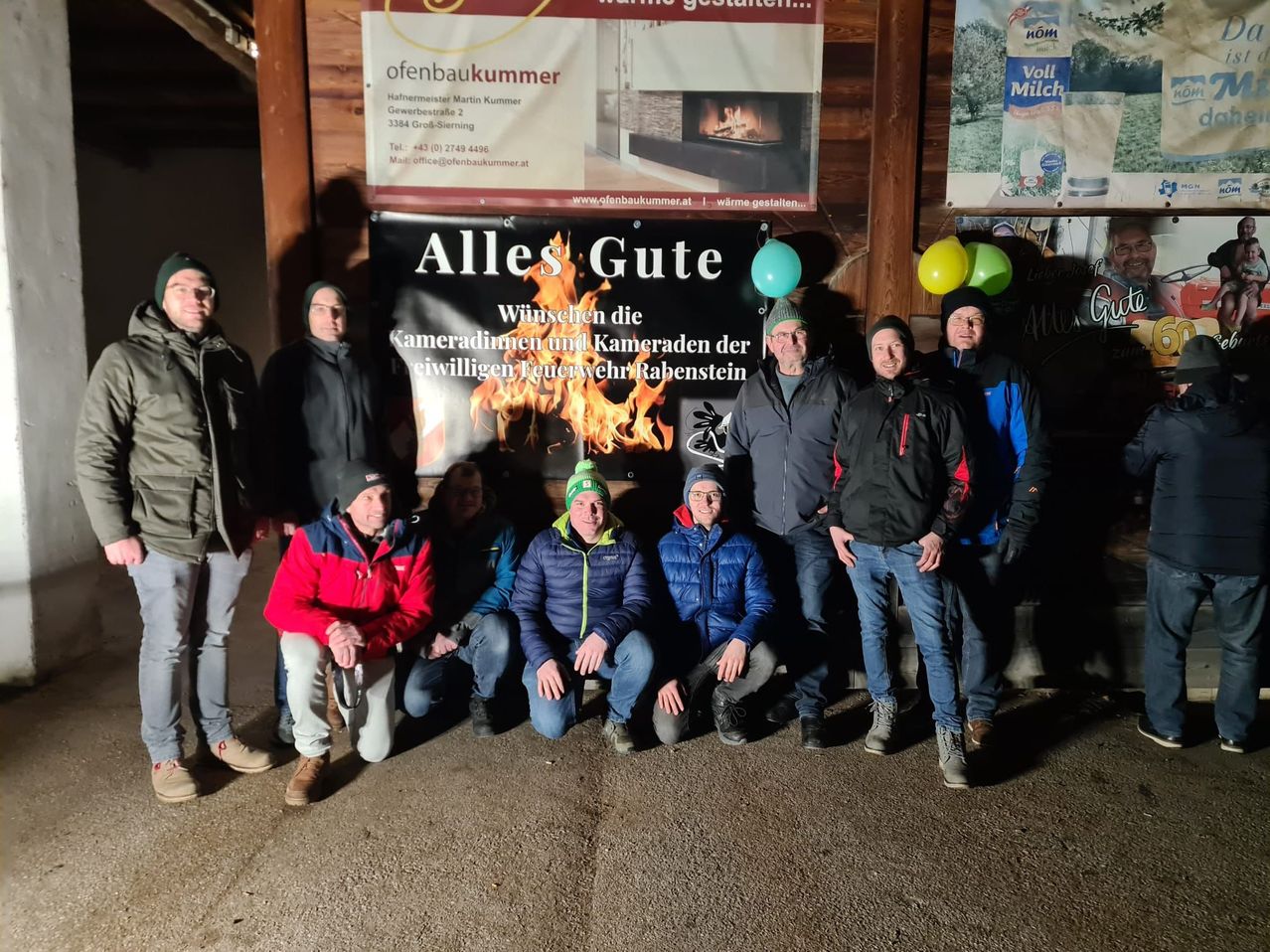 Eine Gruppe von Männern in Winterkleidung posiert für ein Foto vor einem Schild mit der Aufschrift Alles Gute. Dahinter ist ein Kamin zu sehen, und im Hintergrund befinden sich Luftballons.