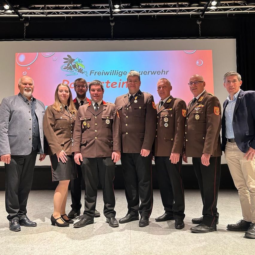 Eine Gruppe von Menschen in Uniformen steht vor einer Bildschirm mit rosa Hintergrund. Sie sind wahrscheinlich Teil einer Zeremonie.