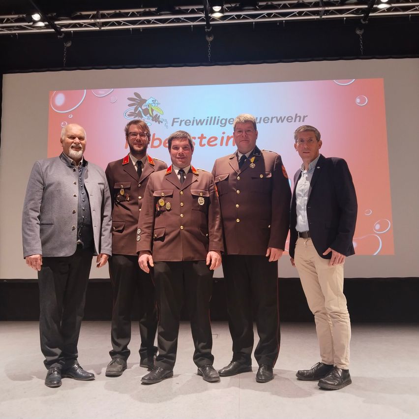 Fünf Männer stehen auf der Bühne, drei in Militäruniformen und zwei in Zivilkleidung, mit einem rosa Hintergrund und Blasen.