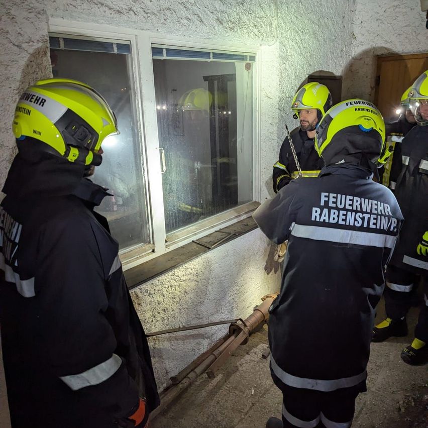 Feuerwehrleute mit gelben Helmen stehen vor einem Glasfenster, möglicherweise bei einer Übung. Sie befinden sich in einem Raum mit weißer Wand und einer Rohrleitung auf dem Boden.