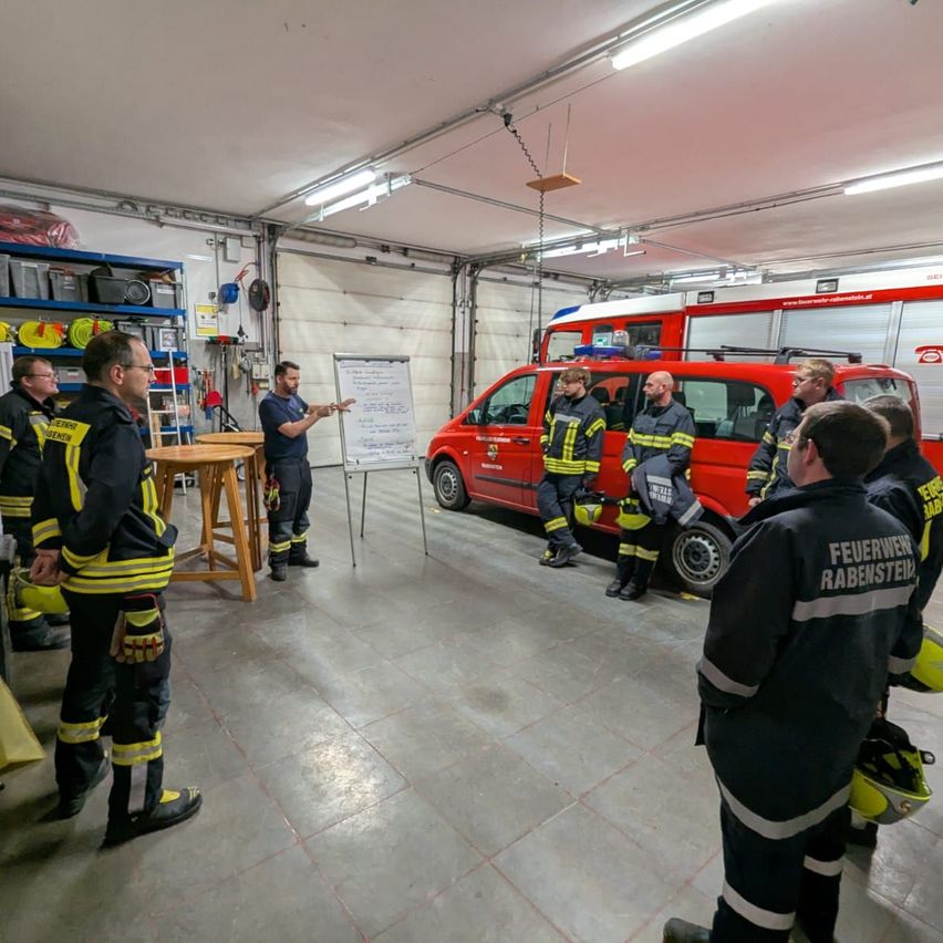 Eine Gruppe von Feuerwehrleuten in Uniform versammelt sich in einer Garage, mit einem roten Feuerwehrwagen in der Mitte. Ein Mann steht vor einer Tafel und hält eine Präsentation.