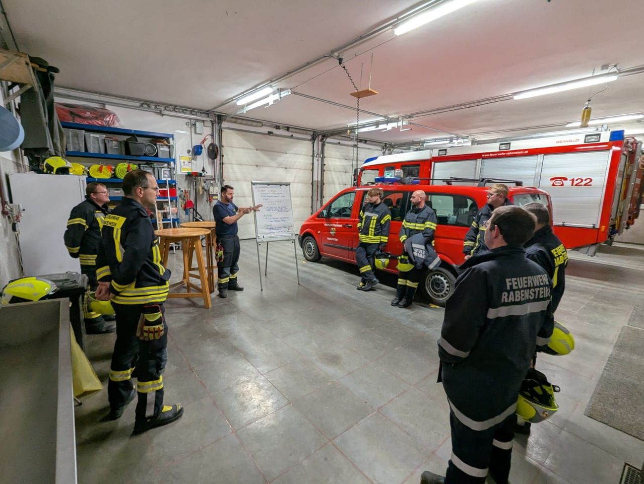 Eine Gruppe von Feuerwehrleuten in Uniform versammelt sich in einer Garage, mit einem roten Feuerwehrwagen in der Mitte. Ein Mann steht vor einer Tafel und hält eine Präsentation.