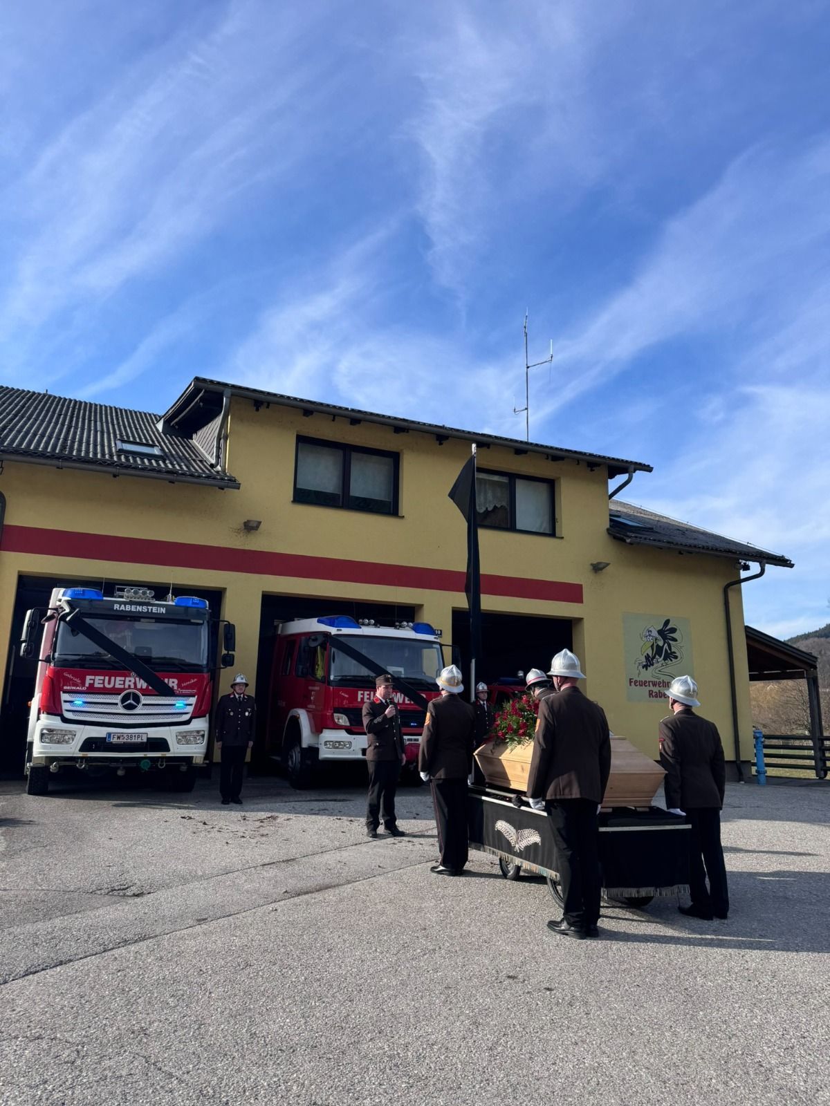 Feuerwehrleute stehen vor einer Feuerwache mit einem Sarg auf einem Karren, umgeben von Blumen. Zwei Feuerwehrwagen sind in der Wache geparkt.